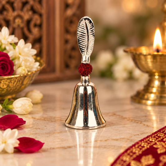 A polished silver-toned metal bell with a decorative leaf-shaped handle and a red cord accent, displayed on a marble surface with flowers, a brass oil lamp, and warm background decor.