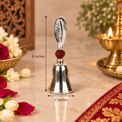 A silver-toned metal bell-shaped ornament with a textured handle and a small dark red decorative accent, shown on a marble surface with dimension arrows indicating 6 inches height and 2 inches width. Decorative flowers, a carved wooden panel, and a brass oil lamp are in the background.