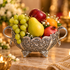 An ornate silver bowl filled with fruit, including green grapes, a yellow pear, an orange, and red apples, set on a marble surface with warm blurred background lights and flowers.