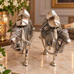 Two ornate silver decorative camel figurines with detailed engravings and draped harnesses, displayed on a polished marble surface in an indoor setting.