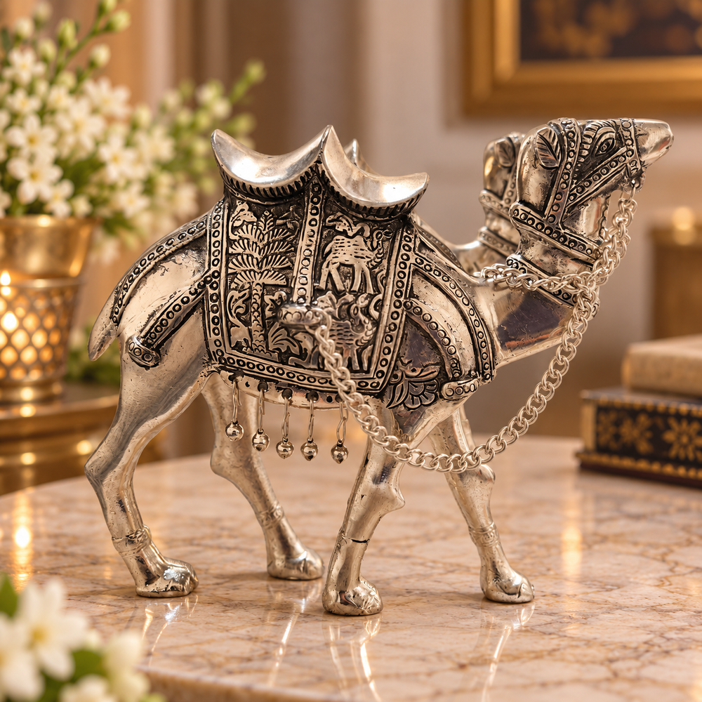 An ornate silver camel figurine with intricate engraved patterns and hanging chain details, displayed on a polished marble surface in a warm, decorated interior setting.