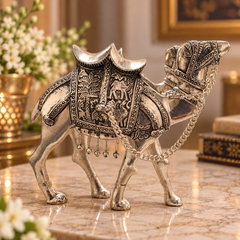 An ornate silver camel figurine with intricate engraved patterns and hanging chain details, displayed on a polished marble surface in a warm, decorated interior setting.