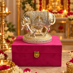 A silver and gold decorative brass Nandi bull statue with a detailed golden harness, displayed on a magenta velvet box in a decorated, warm-toned ritual setting with oil lamps and flowers.