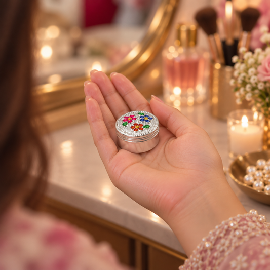 A hand holding a small round silver tin decorated with colorful floral patterns and a rhinestone-embellished lid, set against a softly blurred vanity background with makeup brushes, perfume bottles, and a mirror.