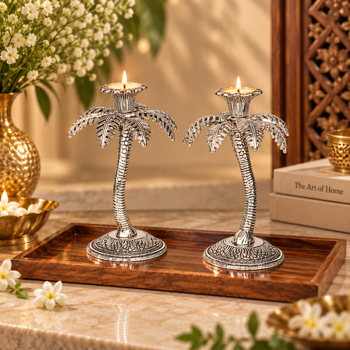 Two silver palm tree candle holders with lit tealights, displayed on a wooden tray on a marble surface with decorative flowers and books in the background.