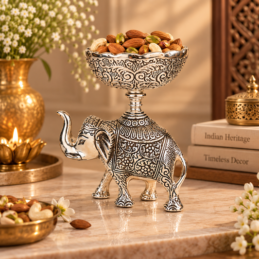 A decorative silver-toned elephant-shaped pedestal bowl filled with almonds and pistachios, styled on a marble surface with gold-toned vessels, flowers, and books in the background.