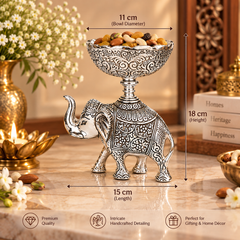 An ornate silver elephant-shaped pedestal bowl filled with nuts, shown on a marble surface with overlaid dimension labels (11 cm bowl diameter, 18 cm height, 15 cm length) and marketing text at the bottom. Decorative flowers, a brass candle holder, and stacked books are in the background.