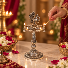 An ornate silver-toned brass oil lamp holder with a peacock finial, lit and set on a marble table. A hand is lighting one of the flames with a match.