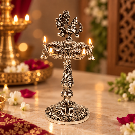 An ornate silver-toned brass oil lamp holder with multiple lit candles, set on a marble table in a decorated indoor setting with warm bokeh lights and floral accents.