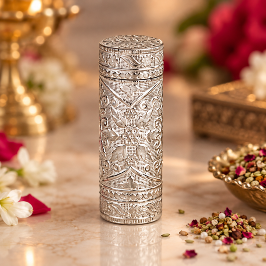 An ornate cylindrical silver-toned metal container with intricate floral engravings, standing on a marble surface with decorative flowers and dried botanicals in a softly blurred setting.