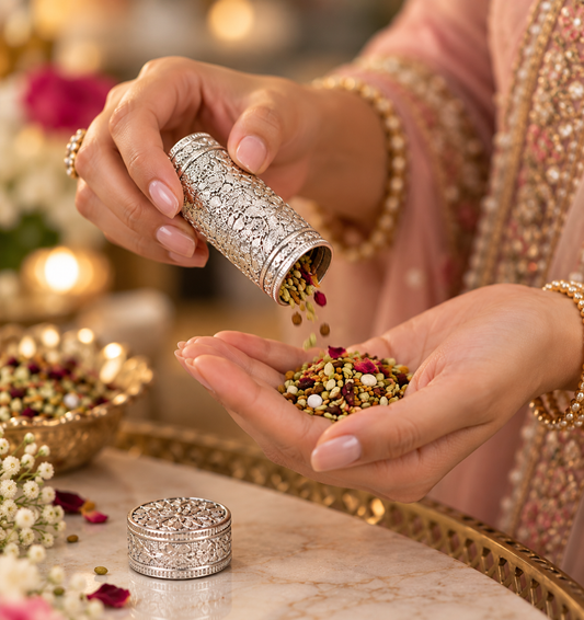 A close-up lifestyle shot of a person pouring dried multicolor lentils from an ornate silver cylindrical container into their hand. The person is wearing a pink embroidered traditional outfit and gold jewelry, with a matching silver container resting on a marble surface in the foreground.