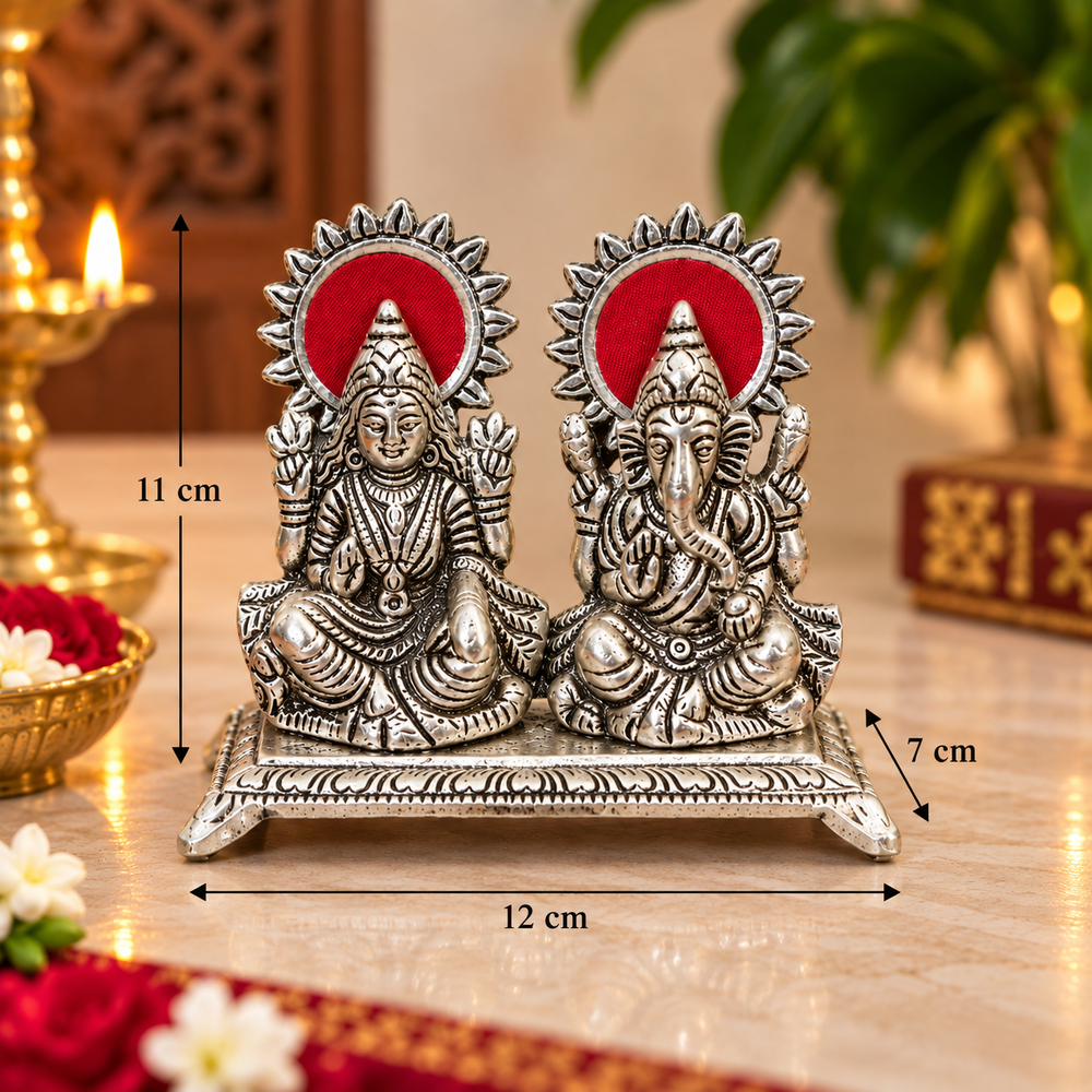 A pair of silver-toned Lord Ganesha and Goddess statue set on a decorative base, shown with overlaid dimension markers indicating 12 cm width, 11 cm height, and 7 cm depth. The background is a warm, softly blurred indoor setting with a lit candle and green foliage.