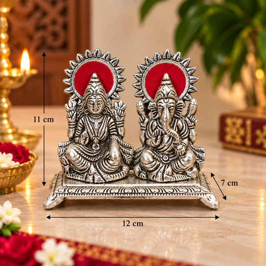 A pair of silver-toned Lord Ganesha and Goddess statue set on a decorative base, shown with overlaid dimension markers indicating 12 cm width, 11 cm height, and 7 cm depth. The background is a warm, softly blurred indoor setting with a lit candle and green foliage.