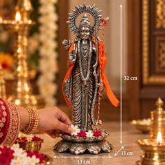 A silver-toned Hindu deity statue on a lotus base, shown in a decorated indoor setting with measurement overlays indicating 32 cm height, 13 cm width, and 13 cm depth. A hand in a red embroidered sleeve offers flowers at the base.