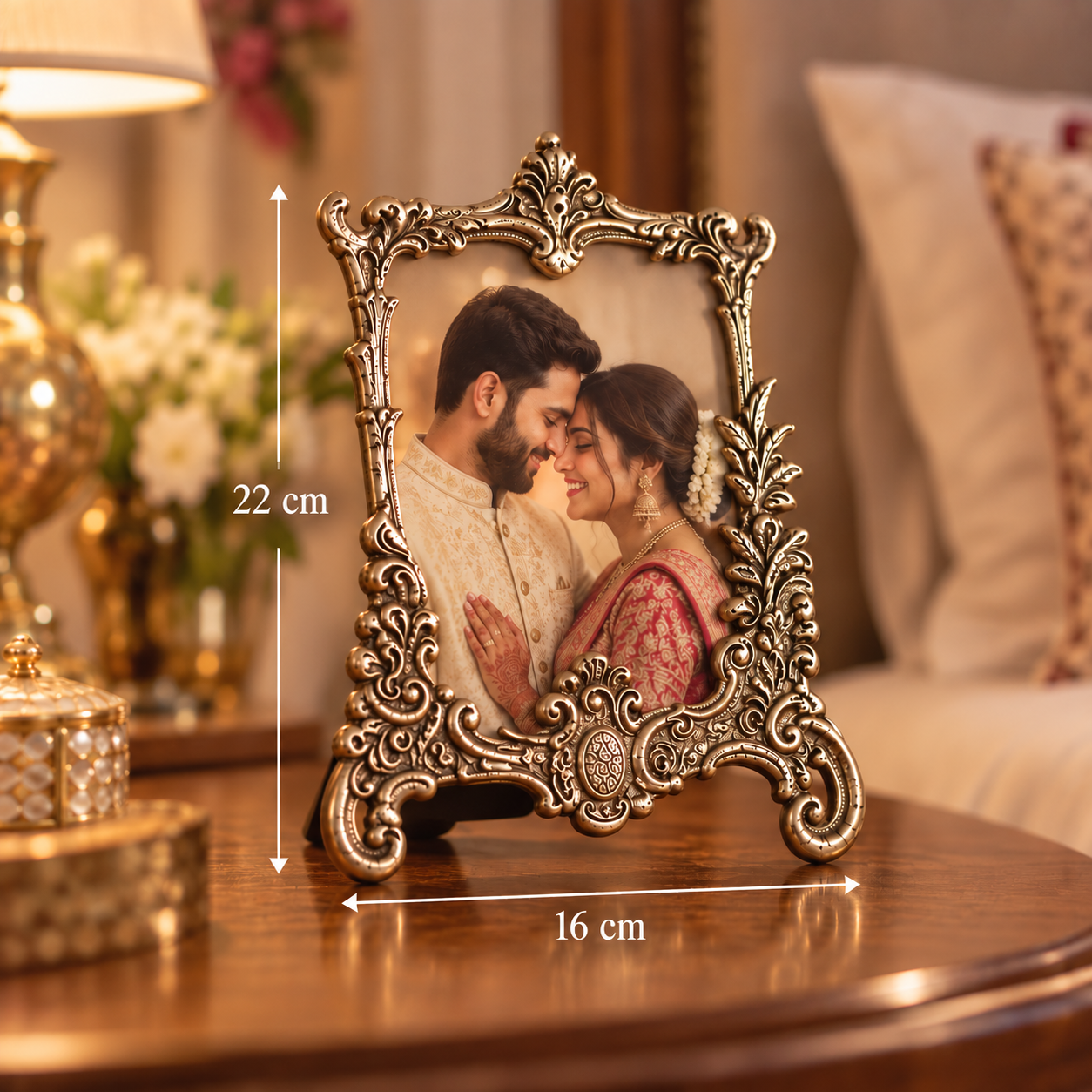 An ornate vintage-style metallic picture frame with a silver-toned finish, holding a photo of a couple. The frame is shown on a wooden table in a warmly lit living room setting, with white dimension lines indicating a height of 22 cm and a base width of 16 cm.