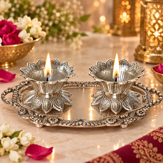 Two silver-toned metal diya lamps with lotus-shaped bases sit on an ornate matching tray on a marble surface, lit with flames, with flowers and warm decorative lanterns in the background.