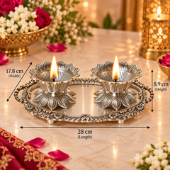 An ornate silver-toned diya holder tray with two lit oil lamps, shown on a marble surface with decorative flowers and warm lights in the background. Black measurement lines and text indicate dimensions: 28 cm length, 17.