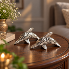 Two silver dolphin figurines with detailed scale textures displayed on a dark wooden table in a cozy, softly lit room.