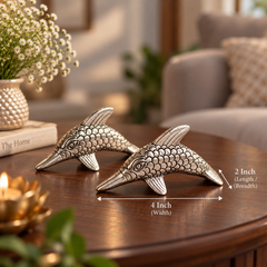 Two metallic dolphin-shaped decorative objects on a wooden table, with overlaid dimension arrows and text showing 4 inch width and 2 inch length/breadth. The scene is a real interior with a sofa, flowers, and candles in the background.