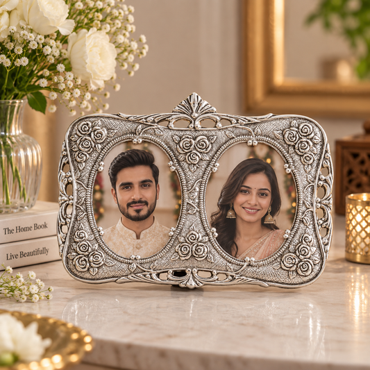 An ornate silver-toned double-photo frame with intricate scrollwork, displayed on a marble surface with a couple's portrait inside. The background features a vase of white flowers, books, and a gold-framed mirror.