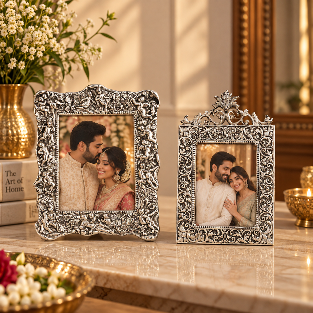 Two ornate silver-toned decorative picture frames with intricate scrollwork, displayed on a marble surface in a warm, elegant interior setting with flowers and candles.