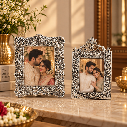 Two ornate silver-toned decorative picture frames with intricate scrollwork, displayed on a marble surface in a warm, elegant interior setting with flowers and candles.