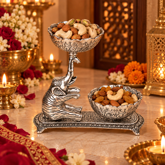An ornate silver-toned elephant-shaped pedestal stand holding two carved bowls filled with nuts and dried fruits, displayed on a marble surface with warm candles, flowers, and decorative lanterns in the background.