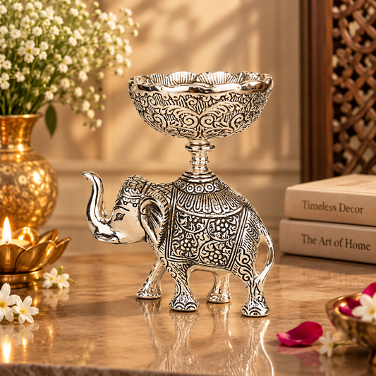 An ornate silver-toned elephant-shaped pedestal candle holder with intricate engraved patterns, displayed on a reflective surface in a warm, decorated interior setting.
