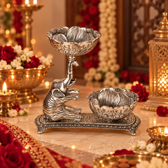 An ornate silver-toned elephant-shaped diya holder with two carved bowls, displayed on a marble surface in a richly decorated festive setting with candles, flowers, and lanterns.