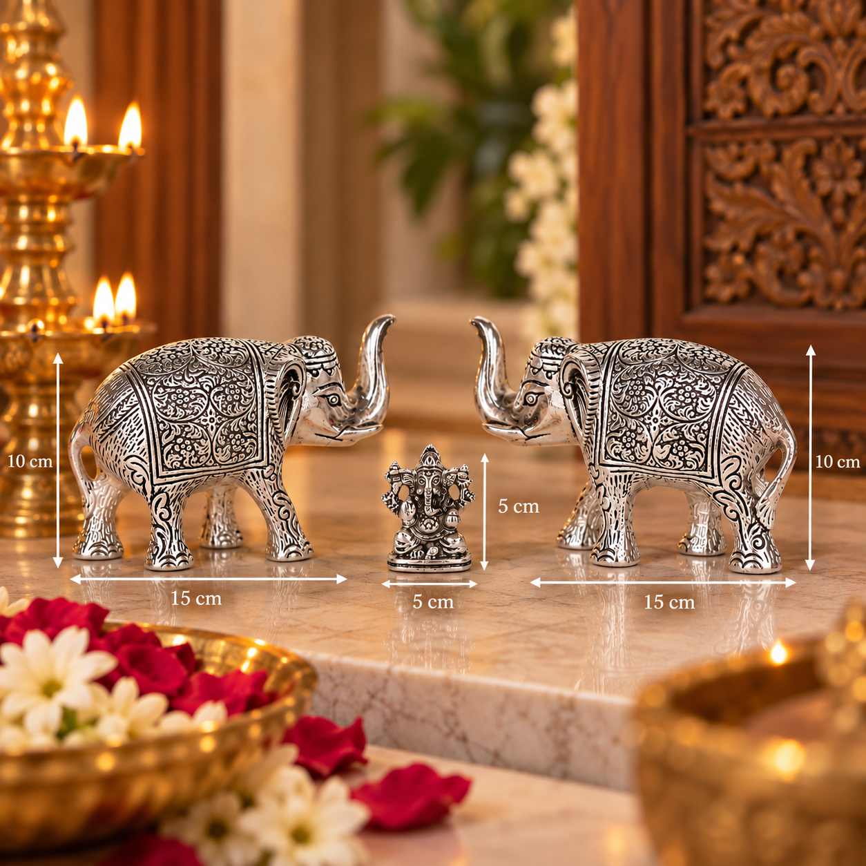 Two ornate silver elephant figurines facing each other with a small central Ganesha statue, shown on a marble surface with overlaid dimension lines indicating 15 cm length and 10 cm height. The scene is styled with lit candles, flowers, and carved wooden decor in the background.