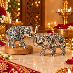 Two ornate silver elephant figurines with intricate embossed patterns, one standing on a round wooden base and the other beside it, displayed on a decorated table with warm lights and floral accents.