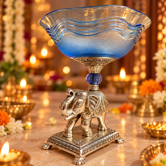 A decorative blue glass and silver elephant pedestal bowl, styled on a marble table with warm glowing candles and floral decorations in the background.