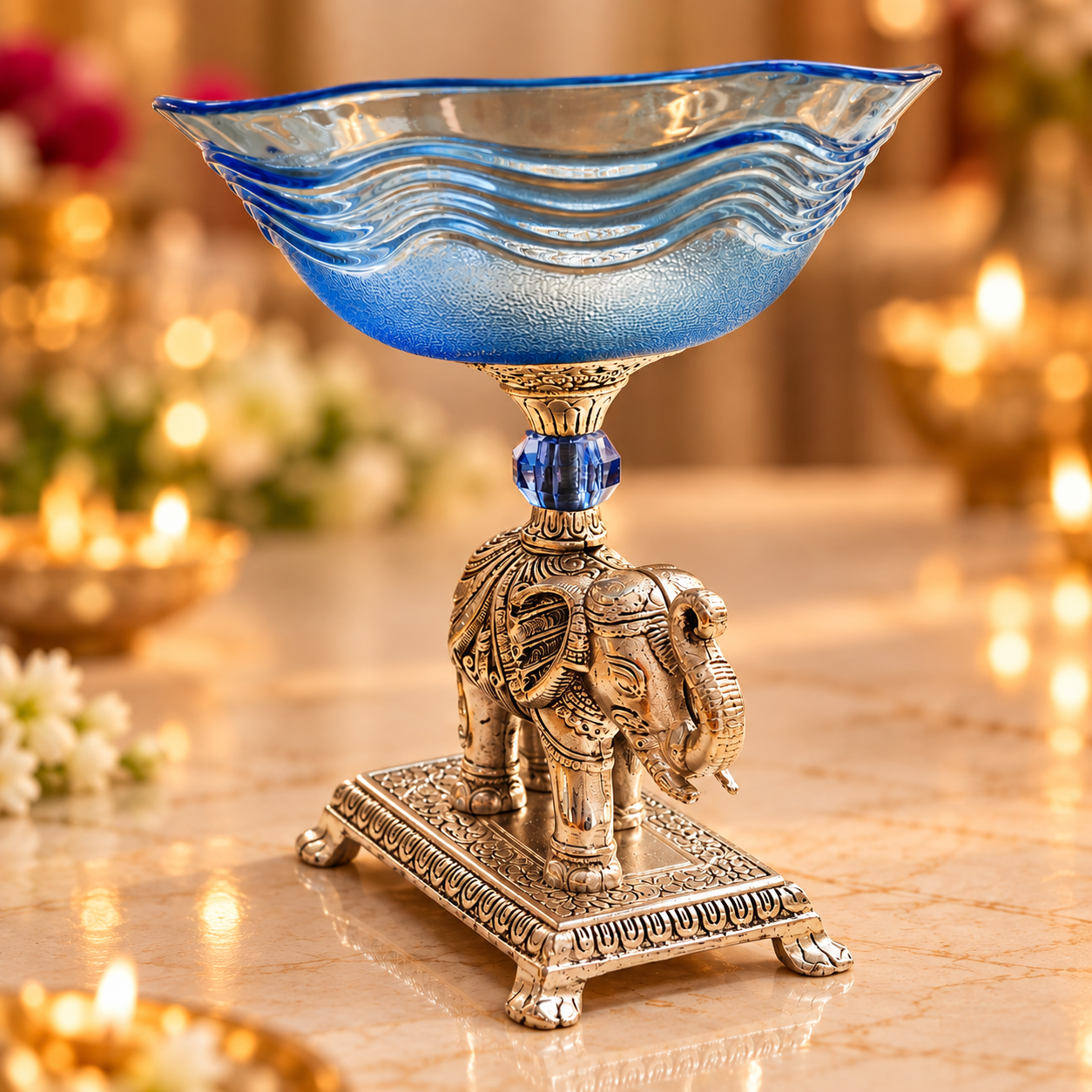 A decorative glass bowl with a wavy blue pattern on a silver-toned elephant pedestal, set on a marble surface with warm blurred background lights.