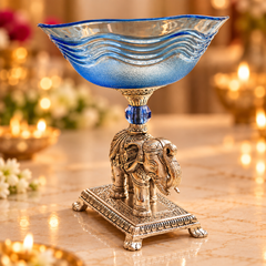 A decorative glass bowl with a wavy blue pattern on a silver-toned elephant pedestal, set on a marble surface with warm blurred background lights.