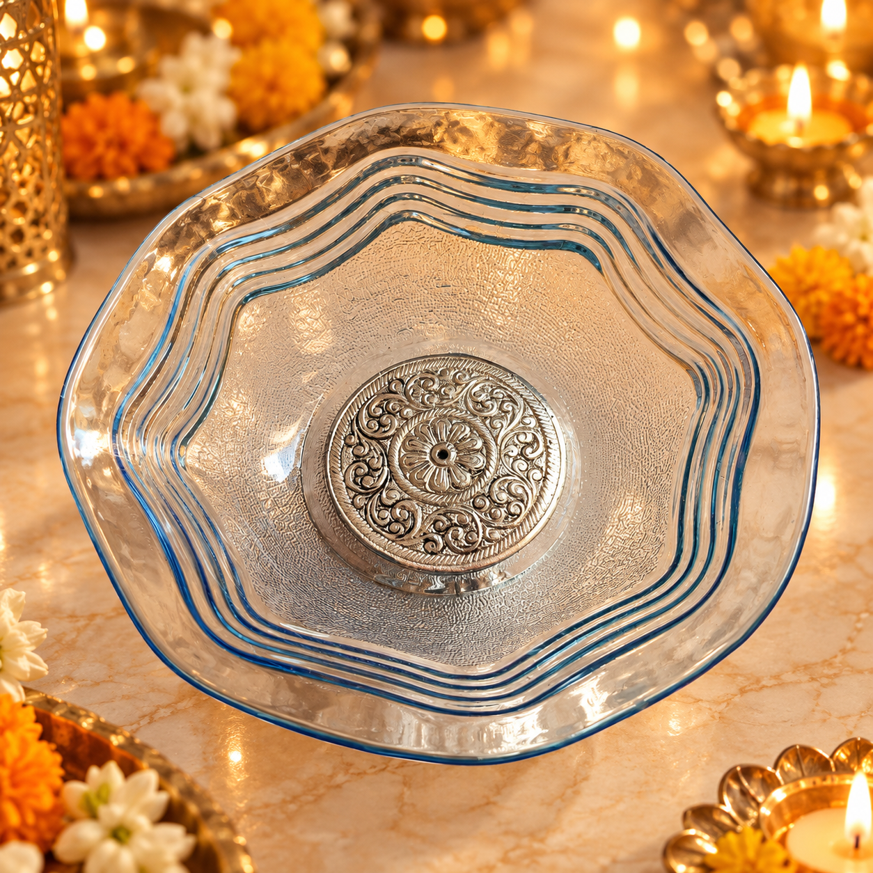 A clear glass bowl with a scalloped rim and blue edging, featuring a raised silver-toned floral medallion in the center, set against a warm, festive background with lit candles and marigold flowers.