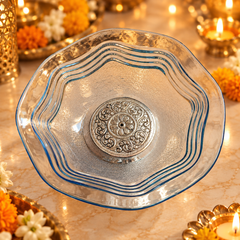 A clear glass bowl with a scalloped rim and blue edging, featuring a raised silver-toned floral medallion in the center, set against a warm, festive background with lit candles and marigold flowers.