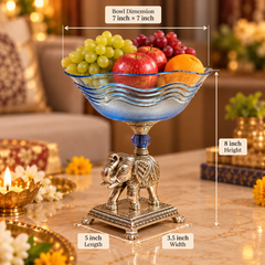 A blue glass fruit bowl with a silver elephant pedestal base, filled with grapes and apples, shown on a marble table with overlaid dimension labels for bowl size, height, length, and width.