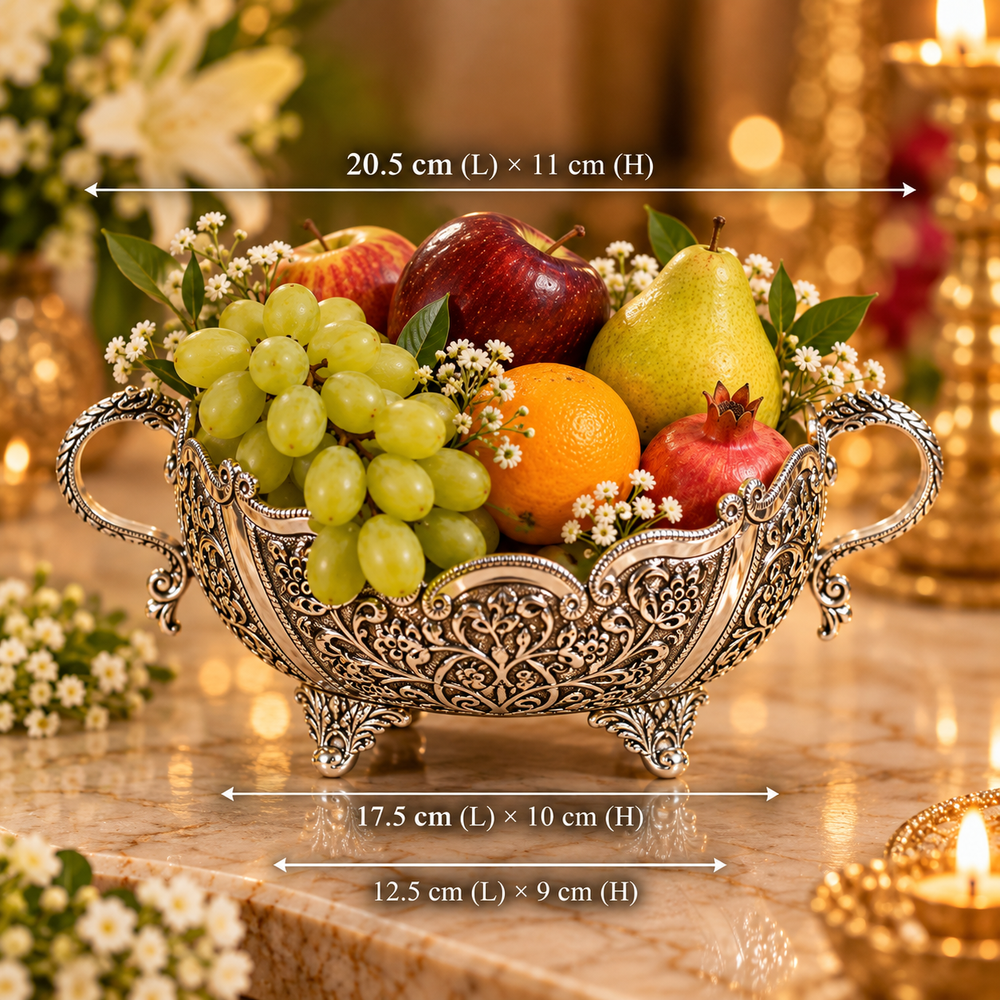 An ornate silver-toned fruit bowl filled with grapes, apple, pear, orange, and pomegranate, shown on a marble surface with overlaid dimension arrows and text indicating its measurements.