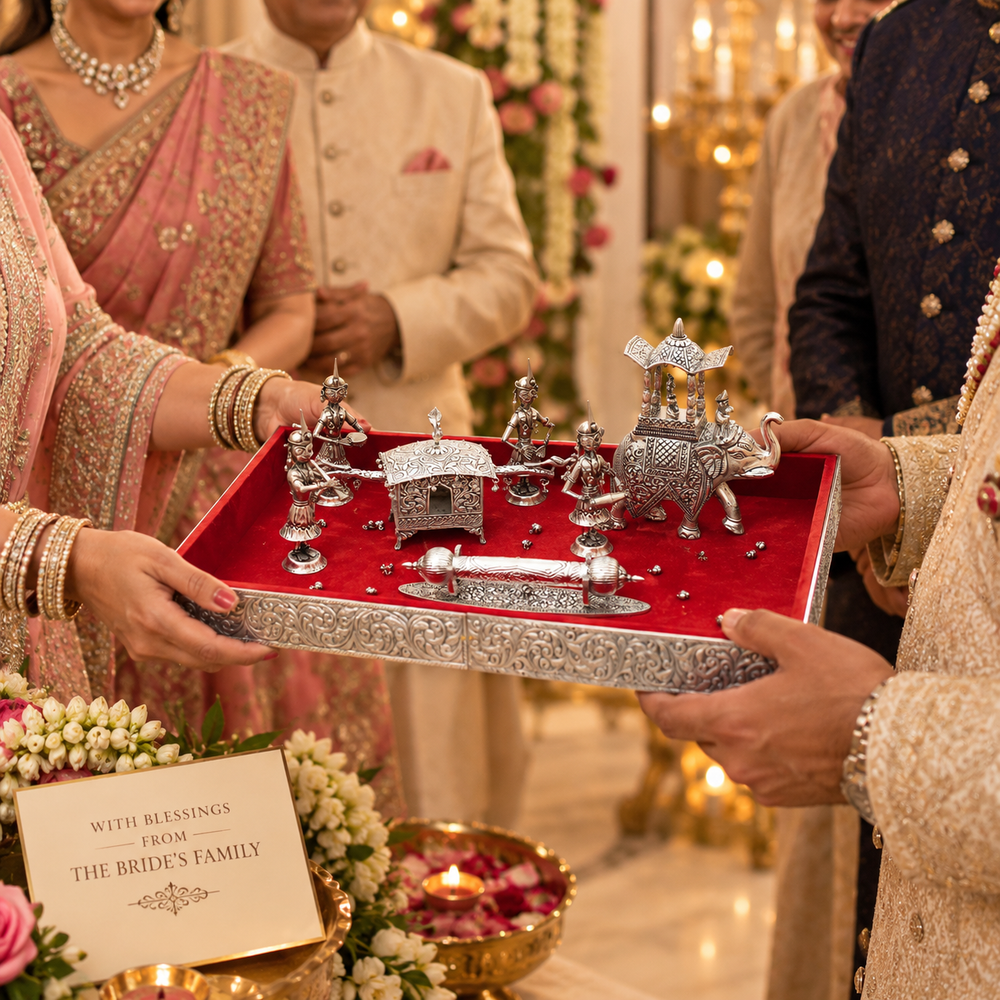 A decorative silver-toned ceremonial tray set with miniature figurines and a red velvet lining is being held by people in traditional Indian wedding attire. The scene includes a bride in a pink saree, a groom in a cream sherwani, and other guests in a warmly lit indoor event setting.
