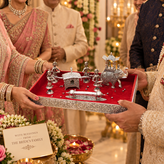 A decorative silver-toned ceremonial tray set with miniature figurines and a red velvet lining is being held by people in traditional Indian wedding attire. The scene includes a bride in a pink saree, a groom in a cream sherwani, and other guests in a warmly lit indoor event setting.