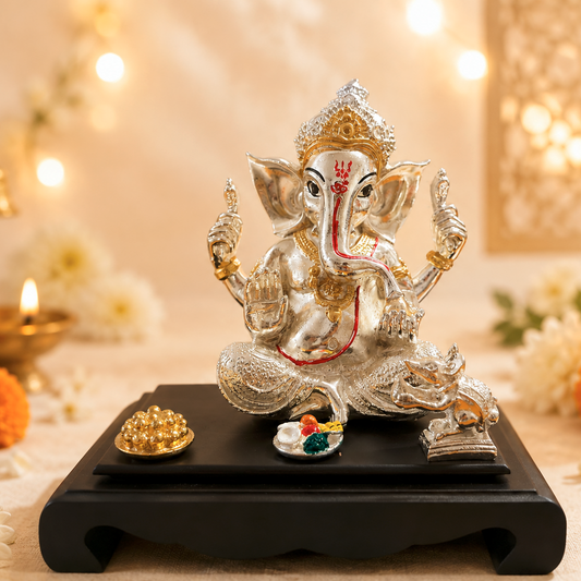 A silver and gold Ganesha statue seated on a black wooden base, decorated with a red tilak and small offerings, surrounded by marigold flowers, white blossoms, and warm bokeh lights in the background.