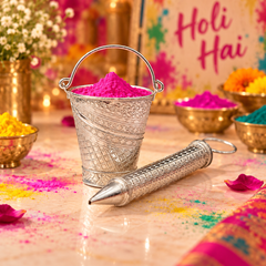 A silver metal Holi pen lies on a marble surface in front of a small silver bucket filled with bright pink powder, with additional bowls of colored powder and a blurred 'Holi Hai' sign in the background.