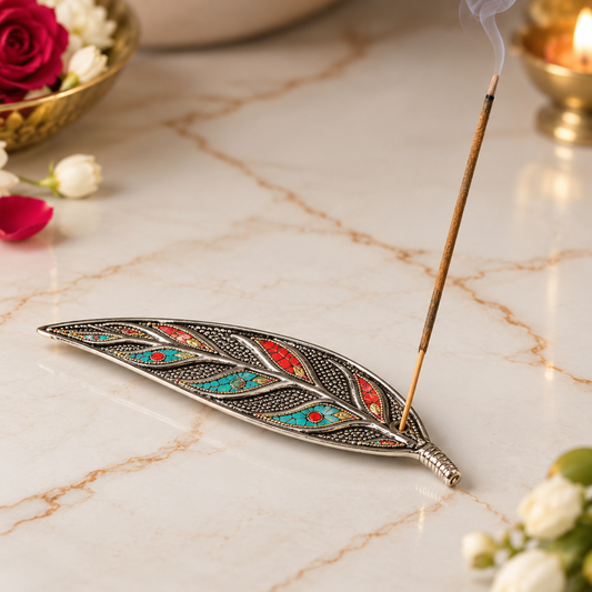 A silver-toned leaf-shaped incense holder with intricate turquoise, red, and gold inlay details, holding a smoking incense stick on a polished cream marble surface with decorative flowers and a candle in the background.