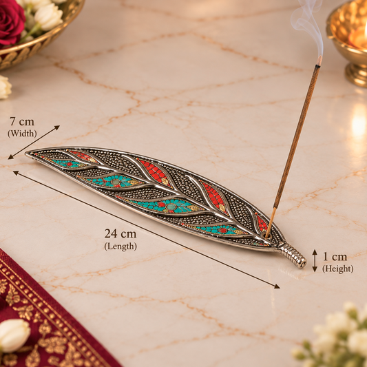 An ornate silver-toned leaf-shaped incense holder with turquoise, red, and gold inlay, shown on a marble surface with a burning incense stick. Measurement arrows and text indicate the product dimensions: 24 cm length, 7 cm width, and 1 cm height.