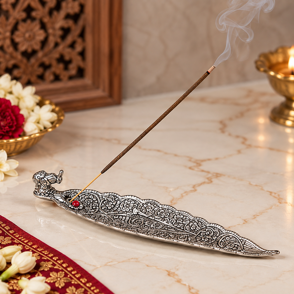 An ornate silver elephant-shaped incense holder on a marble surface, with a smoking incense stick resting across it. Decorative flowers, a carved wooden screen, and a brass lamp are visible in the background.