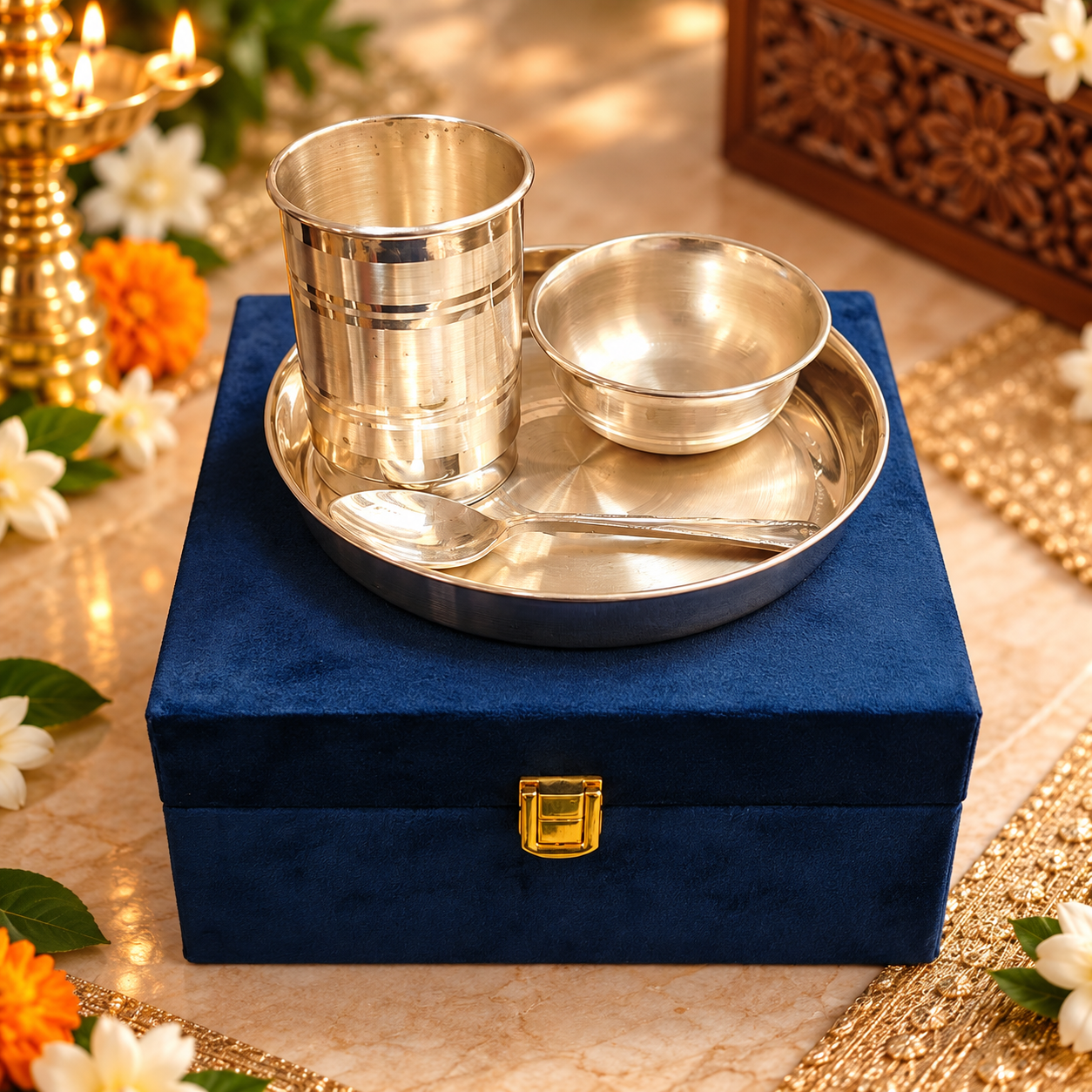 A stainless steel cup, bowl, and spoon set presented on a round silver tray atop a dark blue velvet box with a gold clasp, styled with flowers and warm decorative lighting.