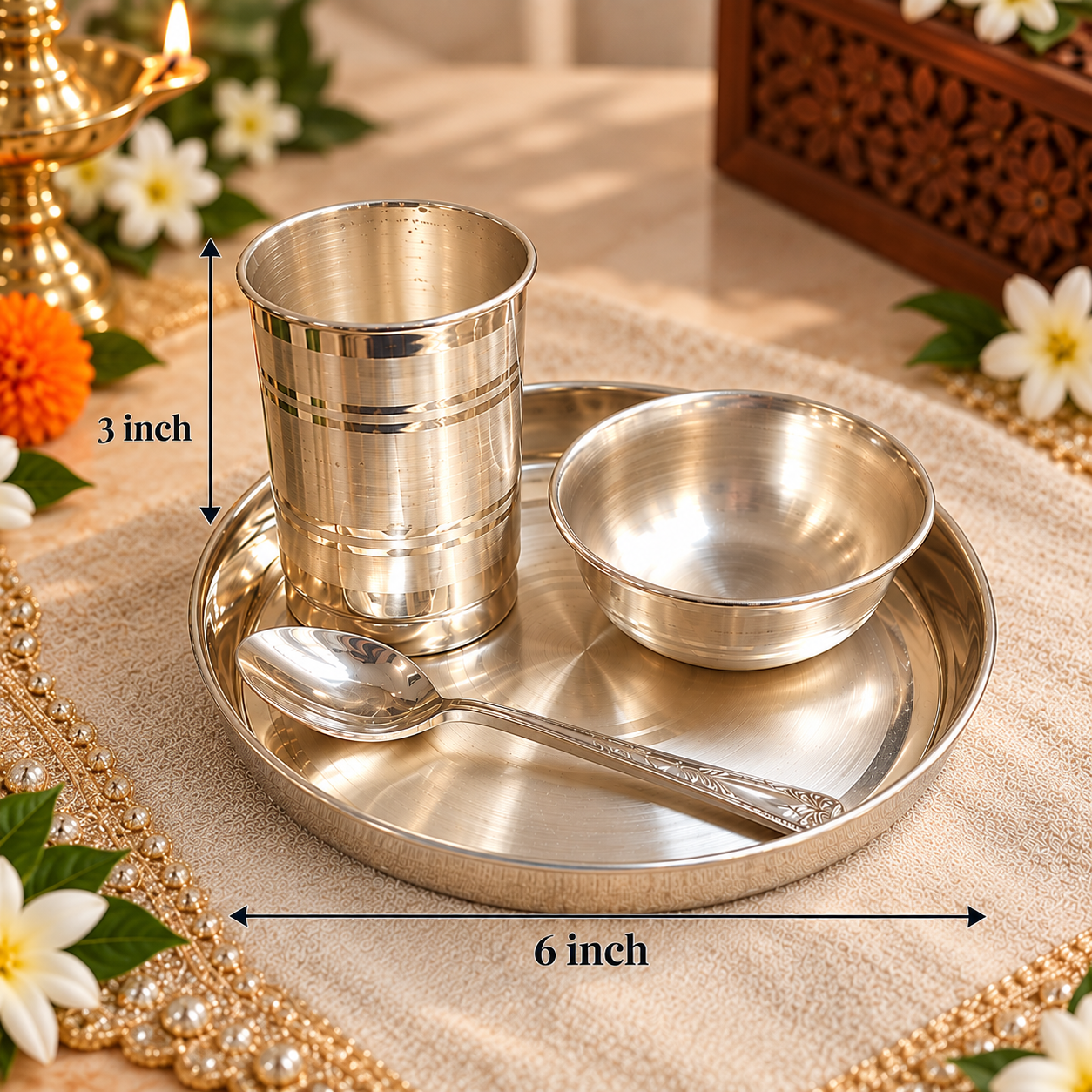 A stainless steel Indian-style prayer tray set with a cup, bowl, and spoon on a round tray, shown with dimension labels indicating 6 inch width and 3 inch height. The tray is placed on a beige fabric with decorative flowers and a brass oil lamp in the background.