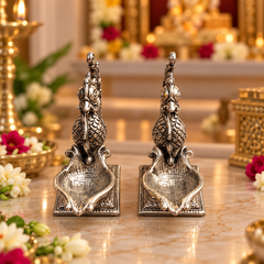 A pair of ornate silver-toned metal peacock-shaped decorative holders or lamps displayed on a polished marble surface, with a softly blurred temple-like interior and flowers in the background.