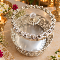 An ornate silver-toned metal storage basket with a floral-patterned handle and a clear glass knob, shown on a decorative tabletop with flowers and warm lighting in the background.