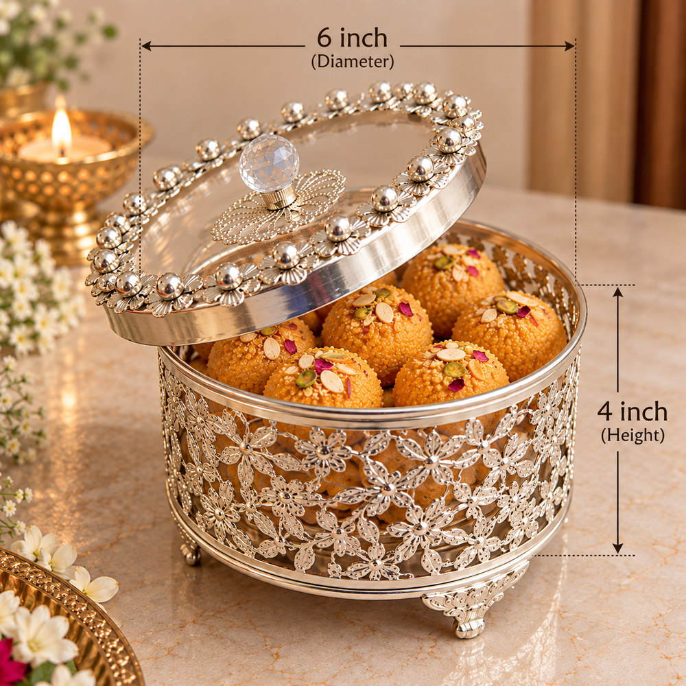A decorative silver-toned metal laddoo box with a floral cutout pattern and a matching embellished lid, shown on a marble surface with sweets inside. Measurement annotations indicate 6 inch diameter and 4 inch height.